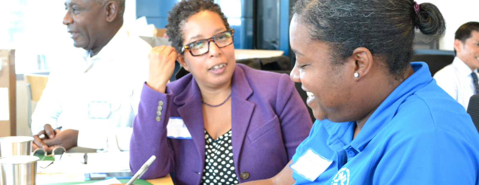 Two women talking at a work event while a man reads something in the background